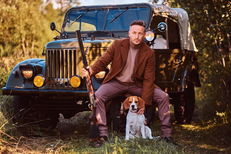 Hunter in a brown coat with a shotgun and a dog beside old-fashioned vehicle in a forest setting— banner for Hunting Travel category, Fieldborne Gear.