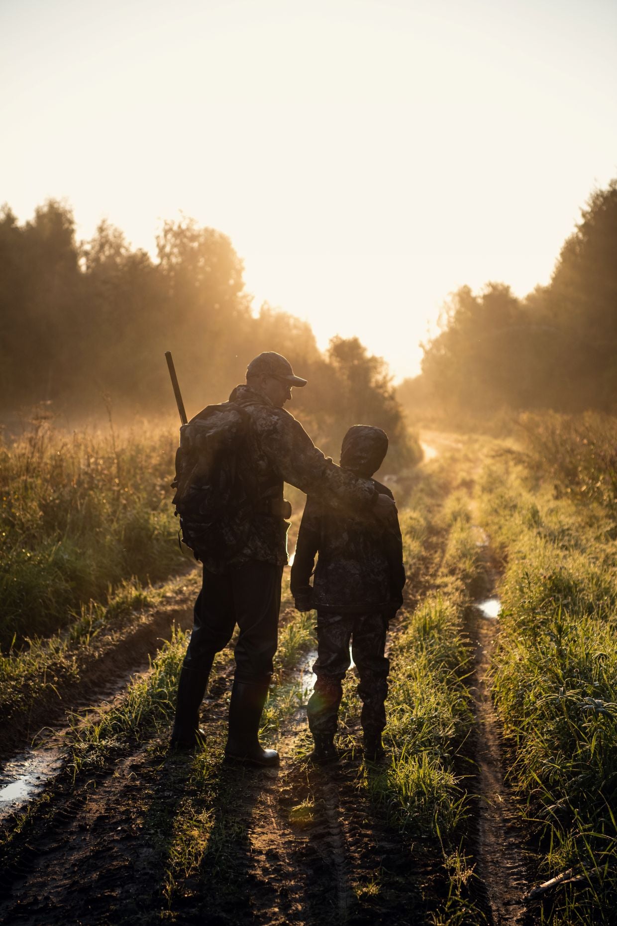 Father and son hunters walking down a forest trail at sunrise — authentic outdoor moment captured in golden light— banner for Stories from the Field category, Fieldborne Gear.